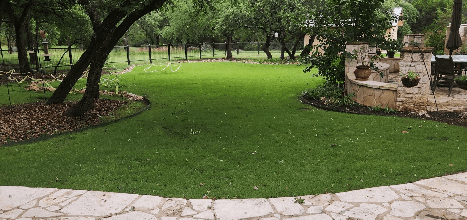 Tree-shaded park lawn with bright green grass, stone edging, and a paved path in front