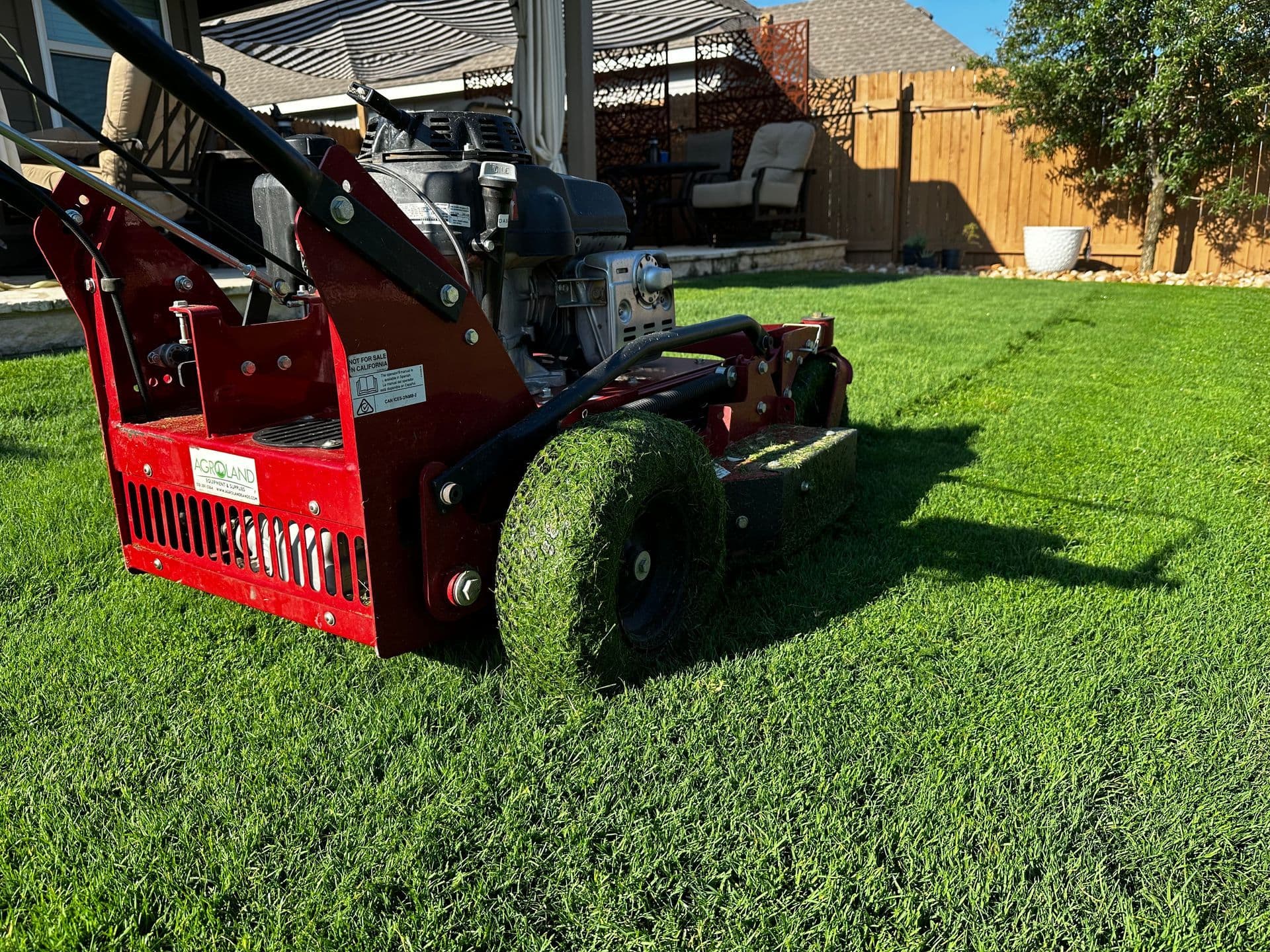 Red walk-behind lawn machine cutting into thick green grass in a fenced backyard.