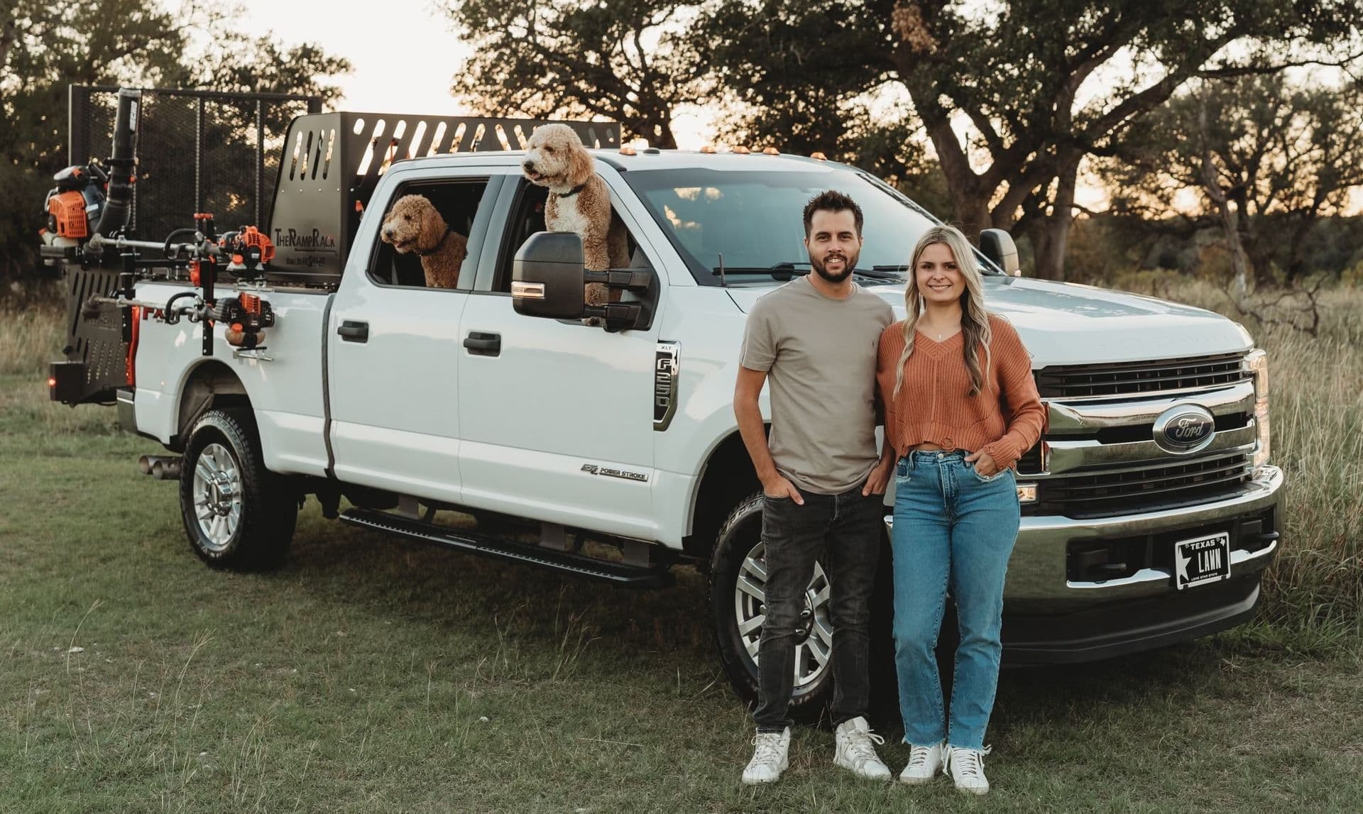 The Local Roots Lawn Maintenance owners beside their white work truck with two golden retrievers.