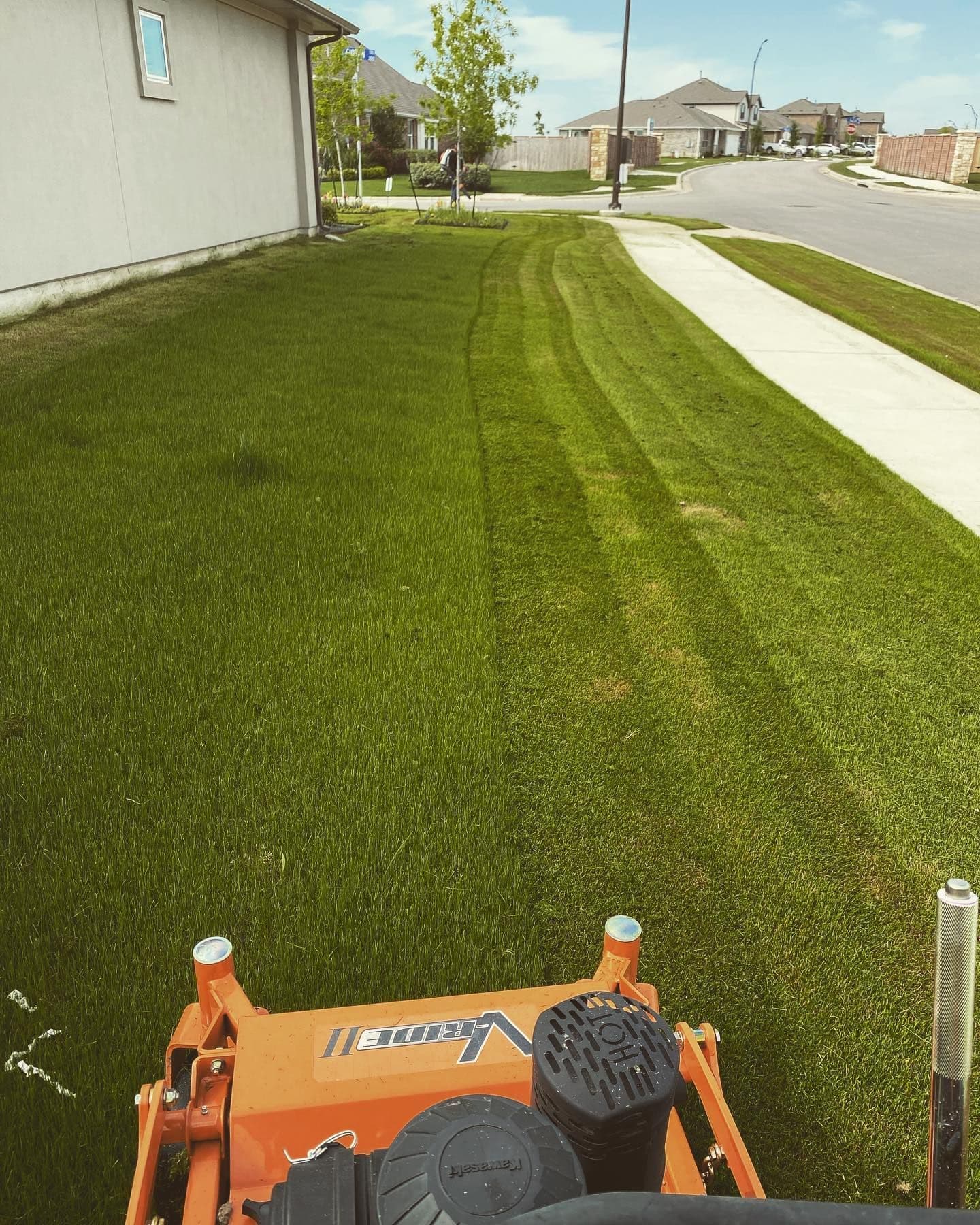 View across an orange mower deck over fresh mowing stripes in a suburban front lawn.