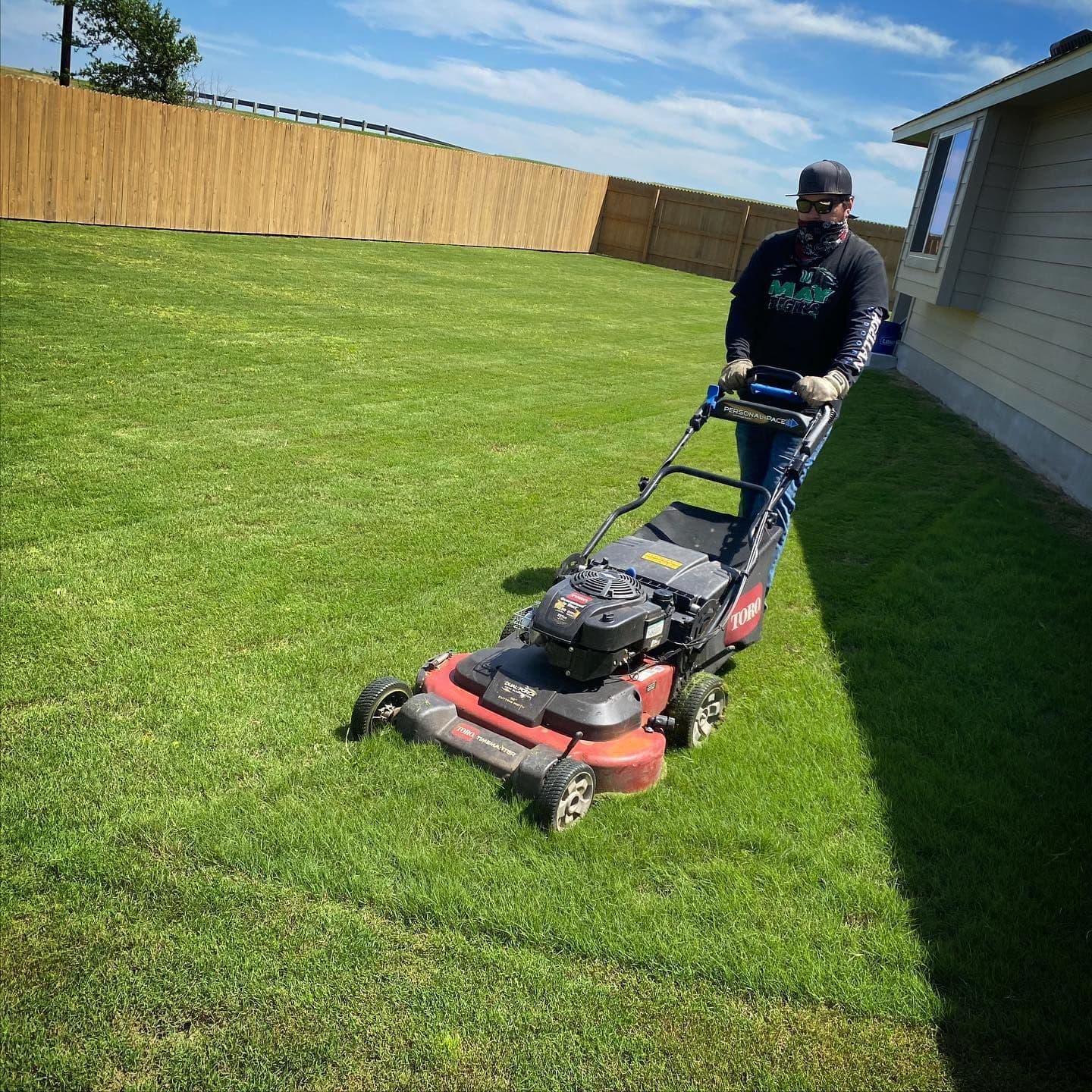 Local Roots crew member mowing a green lawn with a red and black mower beside a wooden fence.