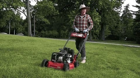 Man in a straw hat mowing a park lawn with a red walk-behind mower.