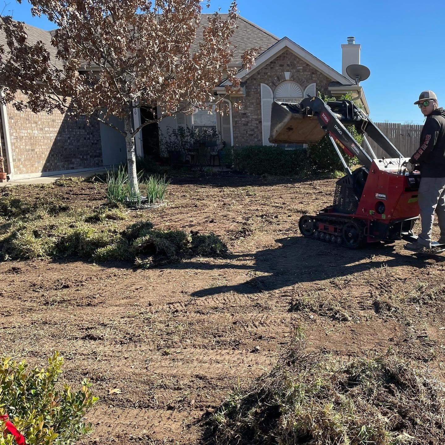 Local roots lawn maintenance worker operating a red mini-excavator in a yard. The ground is dirt and debris near a brick house.