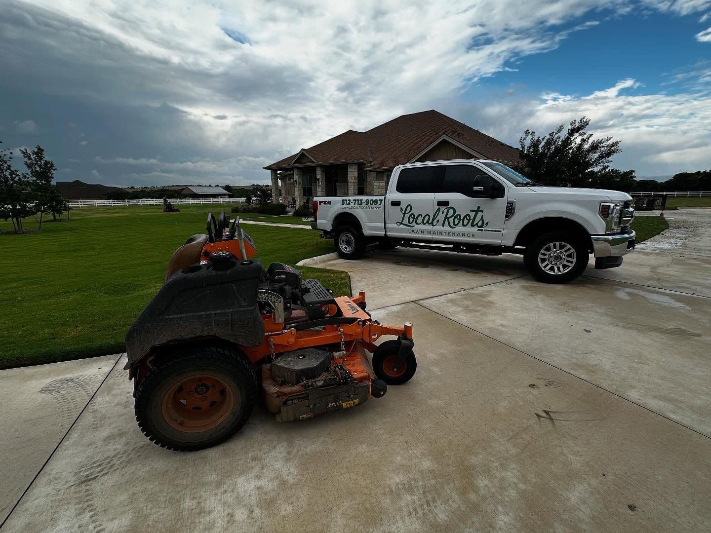 Local Roots work truck and an orange zero-turn mower parked on a driveway beside a freshly mowed lawn.